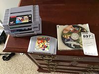 Stack of Super Nintendo game cartridges showing titles including Blazeon, Super Mario Kart, The Lion King, and another cartridge with Spider-Man/X-Men/Arcade's Revenge game label. Nintendo 64 Mario Party cartridge and Rayman Arena PC game CD with auction tag visible.