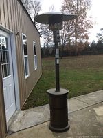 Full view of the propane patio heater standing next to a building with grassy area in the background showing overall height and bent top reflector.