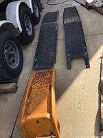 Black metal vehicle ramps side by side on concrete next to trailer tires, showing length and perforated holes.
