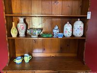 Wide view showing two matching large ceramic canisters with floral design, a floral vase, a vintage Asian bowl, ginger jar, ceramic leaf dish, olive oil bottle with grapes, small candlestick, and sugar and creamer set on a wooden shelf.