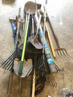 Overall view of yard tools arranged on concrete floor showing variety of axes, shovels, pitchfork, rakes and fabric rolls.