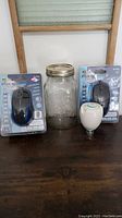 Photo showing two brand new optical mice in packaging, a glass jar with metal lid, and a small white round device on wooden table.