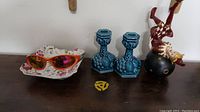 Four assorted items displayed on a dark surface: shell dish with sunglasses, blue candlesticks, clown figurine