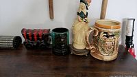 Four ceramic pieces displayed: a red and black covered wagon, green mug, porcelain woman figurine, and large beer stein.