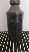 Photo showing front view of dark brown glazed stoneware crock bottle with embossed circular text