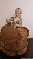 Front view of the porcelain lady figurine with fabric pin cushion, showing detailed facial features, painted rose on chest, and brown fabric base with ruffled trim.