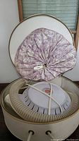 Vintage hair dryer with fabric hood inside round case, flexible hose visible.