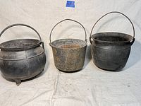 Photo showing three large antique cast iron pots or cauldrons side by side against a white cloth background, each with a metal handle for carrying; one has three legs, one is rusted