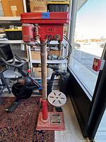 Full view of the red Amrox AF12 floor drill press standing on a cast iron base with round worktable, visible in workshop environment.
