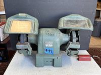 Front view of Black & Decker bench grinder showing dual grinding wheels, protective shields with transparent covers, and blue lot sticker.