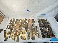 Wide shot of numerous brass backplates and assorted door hardware spread on a white surface with a blue tape label marked '32'. A plastic container holds additional pieces like knobs and locks.
