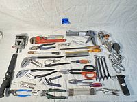 Wide view of all assorted hand tools laid out on white cloth showing variety including pliers, wrenches, clamps, cutting tools, saw, caulking gun, and screwdriver set.