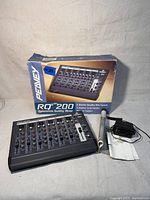 Peavey RQ200 mixer with original box, power supply, and manual rolled up, shown on a surface with gray background.