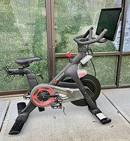 Peloton bike on rubber mat, front-left angle showing touchscreen and resistance knob