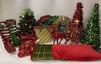 Overall lot showing various Christmas decorations including elf boots, glittery animal figurines, glittered trees and tree toppers, decorative gift bags, and a tree skirt beneath.