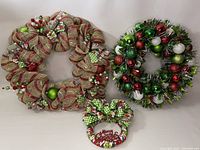 Lot of three assorted Christmas wreaths including burlap ribbon, tinsel with ornaments, and a small wreath with bow and holiday sign.
