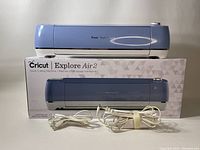 Front view of the Cricut Explore Air 2 machine on its box with power and USB cords in front.