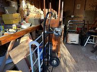 Black metal heavy duty hand truck with two large wheels, one damaged needing replacement, placed amidst workshop clutter with other items in background.
