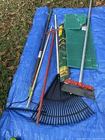 Photo showing multiple garden tools on blue tarpaulin on grass, including metal hoe, black plastic leaf rake, metal leaf rake, red hand tool, green kneeling pads, and rolled green lawn edging strip.