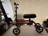 Side view of pink knee scooter showing the black cushioned knee pad, black foam handlebar grips, pink frame, and four black wheels, with basket visible in the background.