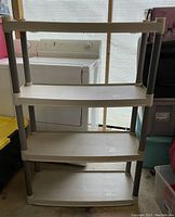 Photo of a four-tier plastic shelving unit with white shelves and grey legs placed in a garage or laundry area.