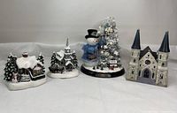 Four Christmas-themed collectibles, including cottages, a church, and snowman with tree, all snowy and detailed.