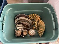 Top-down view of a plastic storage tote containing various vintage metal trays, brass folding hand fans, and small lidded sugar bowl.