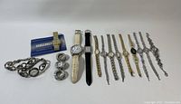 Wide view of all eleven watches laid out in a row with two small circular metal accessories and Seiko manual on left in white background.