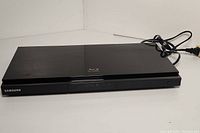 Photo showing front left angle of black Samsung BD-D5700 Blu-ray Disc player with power cord visible.