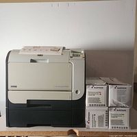 Front view of HP Color LaserJet CP2020 printer with four boxes of Itamx toner cartridges stacked beside it.
