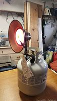 Infrared heater connected on top of a white propane tank, showing the heating element with the orange reflector lit up.