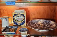 Photo of the blue and white plate, square trinket dish, and porcelain tea canister with decorative medallion, placed on a wooden shelf.