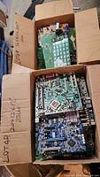 Two open cardboard boxes filled with multiple scrap computer motherboards stacked inside, showing various electronic components and circuitry.