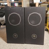 Front view of two stereo speakers with black grilles showing woofer and tweeter outlines and JBL branding on top.