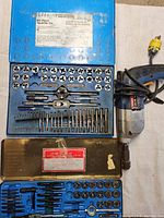 Blue metal case open showing 60-piece tap and die set with various round dies, threading handles, and taps; electric tapper drill on the side.