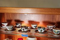 Row of eight vintage tea cup and saucer sets with various Scottish tartan and soldier illustrations, displayed on a wooden shelf.