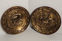 Photo showing two vintage brass wall plates with sailing ships and ornate borders resting against a white background.