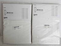 Two unopened packs of Ikea Hilja curtains in white, each pack containing two curtains, seen folded and sealed in original packaging.