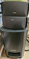 Photo of two compact Bose satellite speakers stacked on top of a large Bose subwoofer, all black with Bose logos visible