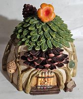 Front view of Hallmark fairy garden house figurine showing pinecone roof, orange flower, tree branch walls, and wooden door with steps.