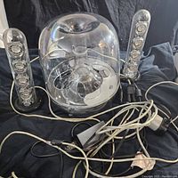 Front view of SoundSticks III multimedia speaker system showing transparent subwoofer and two satellite speakers along with cables and power adapter on black cloth.