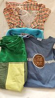 Four assorted men's shirts laid out, including a checkered shirt, blue Andrew Peterson t-shirt, turquoise shirt, and green graphic tee.