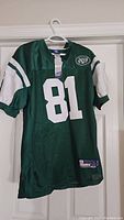 Photo of front side of green NY Jets football jersey with number 81 and team logo patch on chest, displayed on white hanger.
