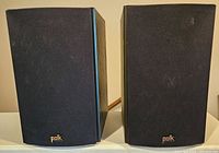 Front view of the pair of Polk T-15 bookshelf speakers with black fabric grilles and Polk logo at bottom.