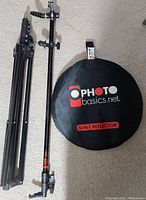 Photo shows folded metal light stand, metal reflector holder, and black carrying case for reflector with Photo Basics branding