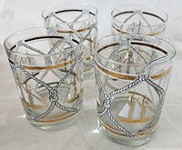 Set of four clear glass drinking glasses with gold, black, and white nautical boat and rope designs arranged side by side on a flat surface.
