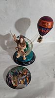 Photo showing Cowardly Lion figure inside clear dome on black base with loose round button featuring Wizard of Oz characters in balloon below, and separate small hot air balloon decoration