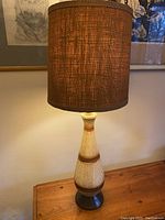 Full view of the lamp showing tall textured base with vertical lined panels and brown burlap cylindrical shade lit, placed on a wooden surface in front of wall art.