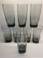 Set of 8 smoky grey glass tumblers arranged three on top row and five on bottom. Tumblers have thick dark grey bases and gently rounded square edges.