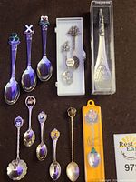Collector spoons displayed on dark surface including a fine pewter Canadian indigenous totem spoon, Alaska totem spoon, Scotland, Wales, Ireland, Jasper, Bermuda, and US state spoons, some in packaging and with a Coca-Cola branded spoon.